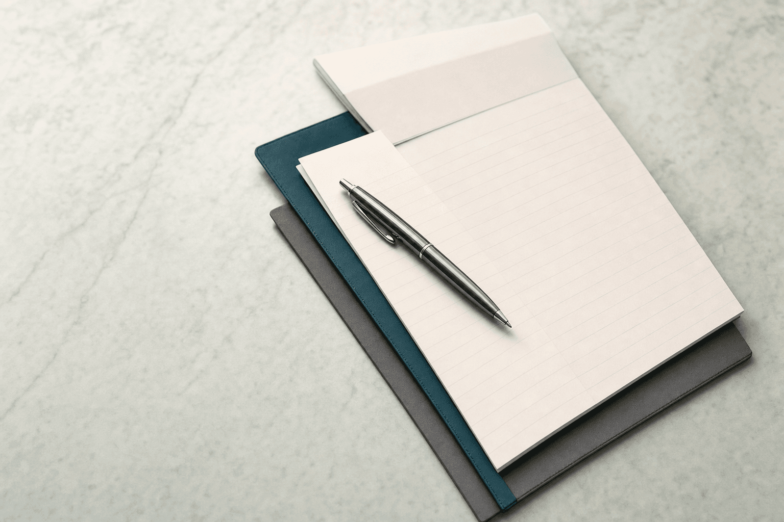 Representative photograph of a blank notepad, teal notebook, and metal pen resting on a marble desk in soft daylight.