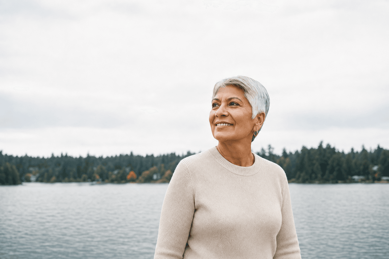 Representative photograph of an older woman standing beside a calm lake with a tree line behind her